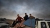 A man and two boys sit on belongings at the back of a truck as they flee Ras al-Ayn, Syria, Oct. 9, 2019, with smoke billowing in the background during a Turkish offensive.
